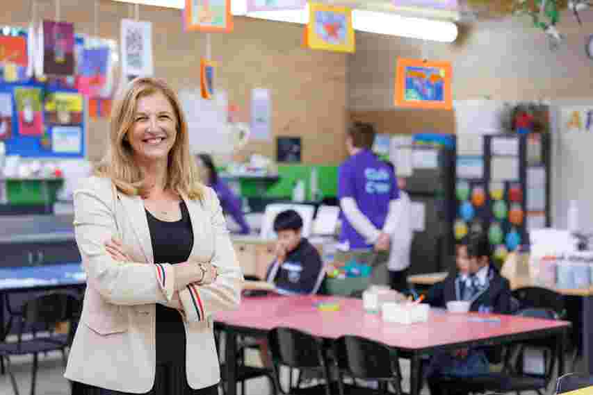 Smiling woman in a beige blazer stands in a colorful classroom with children working at tables in the background.