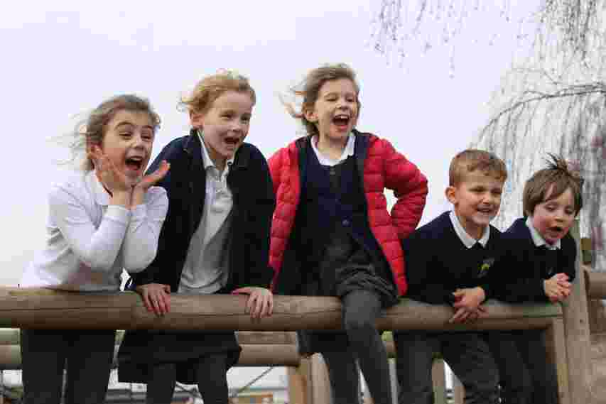 Five children leaning over a wooden railing, smiling and cheering on a windy day.