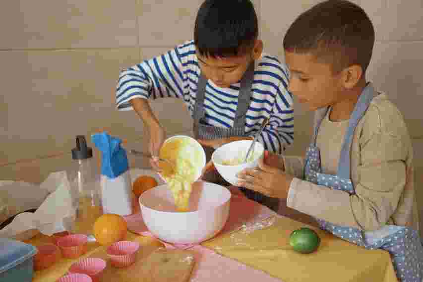 Two boys baking together, pouring batter into a bowl. They wear aprons, surrounded by baking items and colorful cupcake liners.
