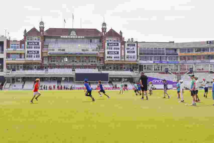 Children playing cricket on a grassy field in front of a large stadium, with coaches and equipment visible in the background.