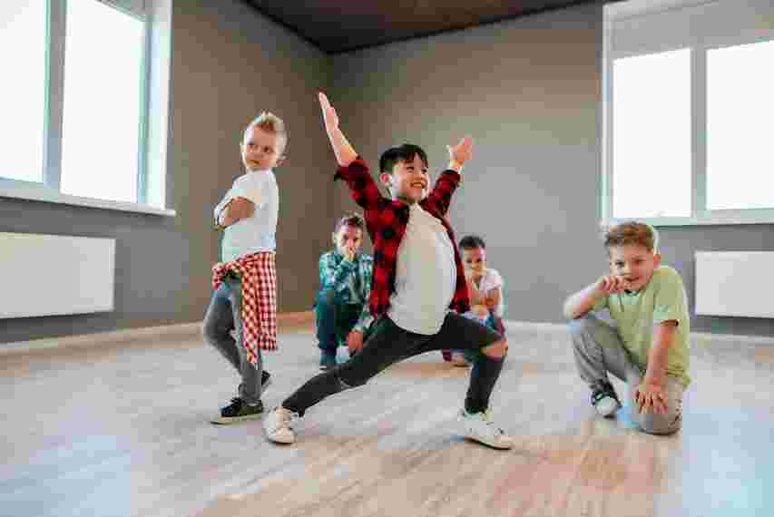 Children are enthusiastically posing and dancing in a spacious room with large windows and wooden floors.