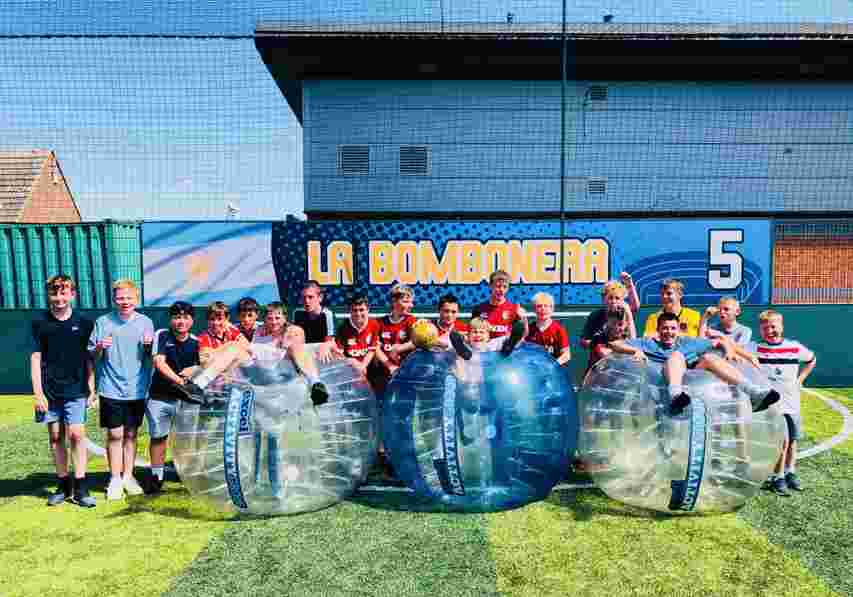 Group of boys on artificial turf posing with three large transparent bumper balls in front of a "La Bombonera" sign.