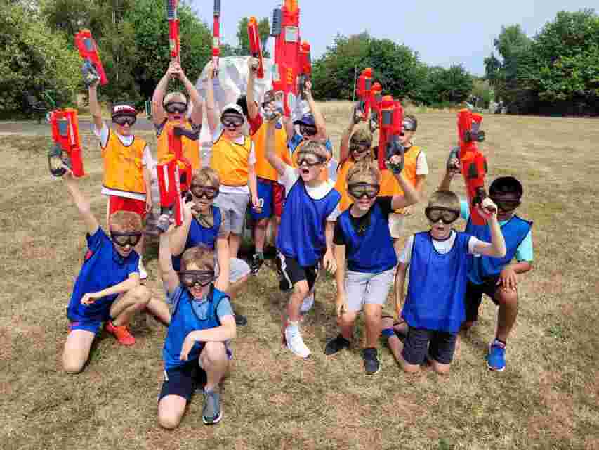 Group of children in blue and orange vests and safety goggles cheering and raising red toy blasters on a dry grassy field.