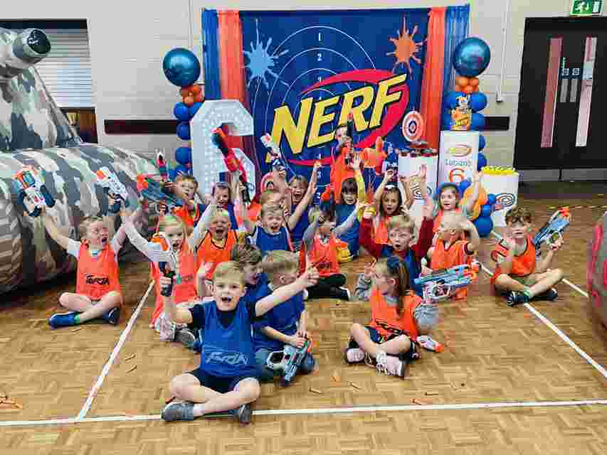Children in orange and blue vests holding Nerf blasters, cheering in front of a colorful Nerf party backdrop.