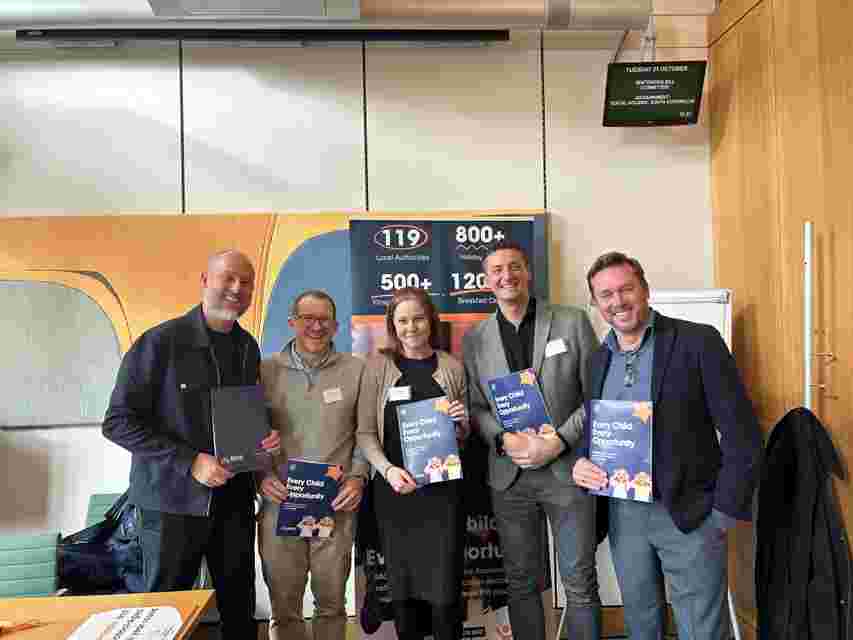Five people smiling and holding booklets titled "Every Child Opportunity" in a conference room with a banner and screen in the background.