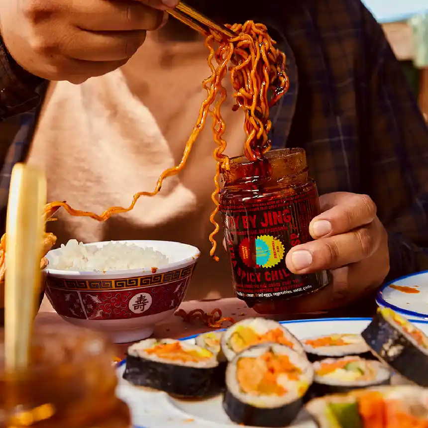 Noodles being dipped into a jar of Xtra Spicy Chili Crisp at a table, surrounded by plates of food