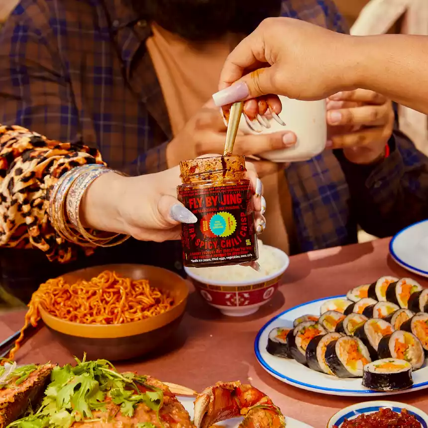 A person's hand holding a jar of Xtra Spicy Chili Crisp at a table, surrounded by food and other hands