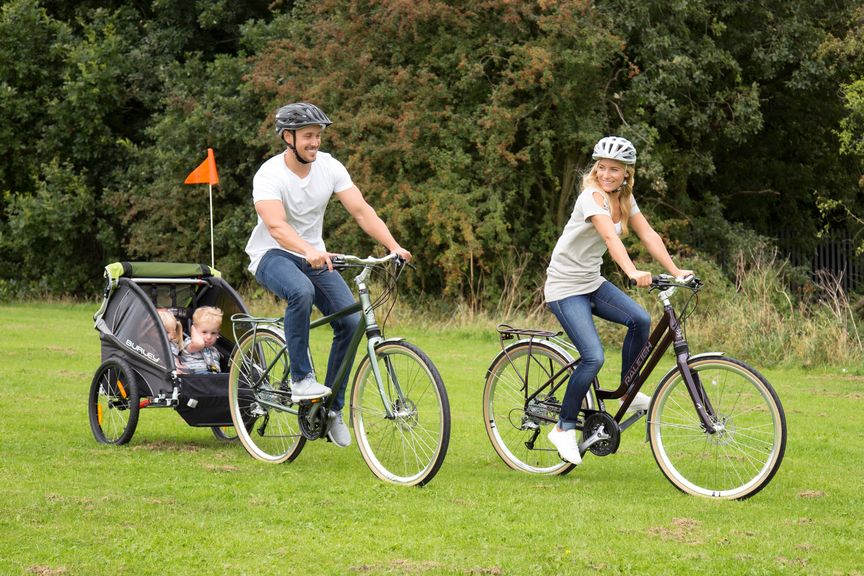 Two adults on a bike with a bike trailer and kids