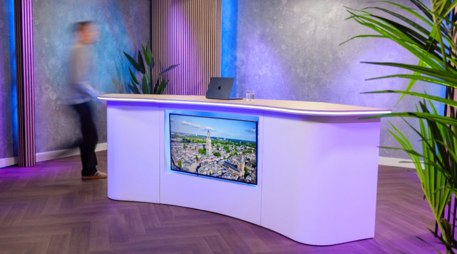 Modern studio setup with a curved white desk, laptop, and cityscape screen; a blurred person walks past, with vibrant purple and blue lighting.