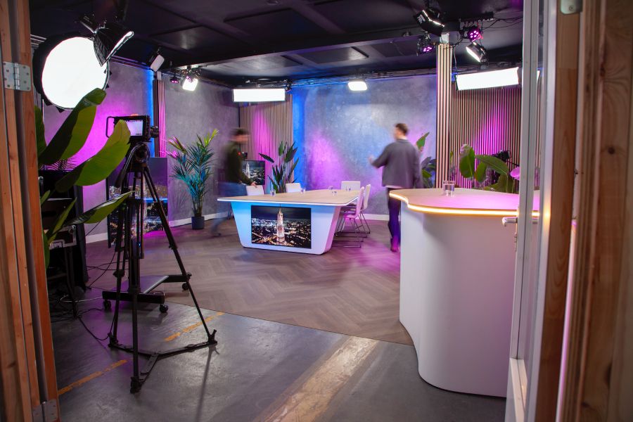 A modern studio with a table, lighting equipment, and plants. Two blurred figures are present, and the lighting casts purple and blue hues.