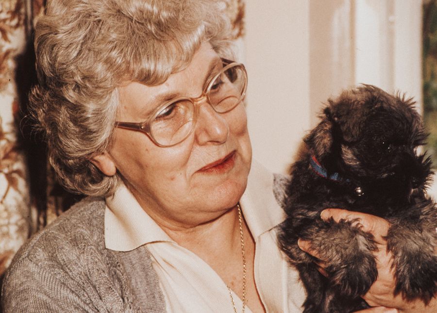 SRS care- photo of an older lady with curly grey hair and grey cardigan holding a small brown Maltese type puppy