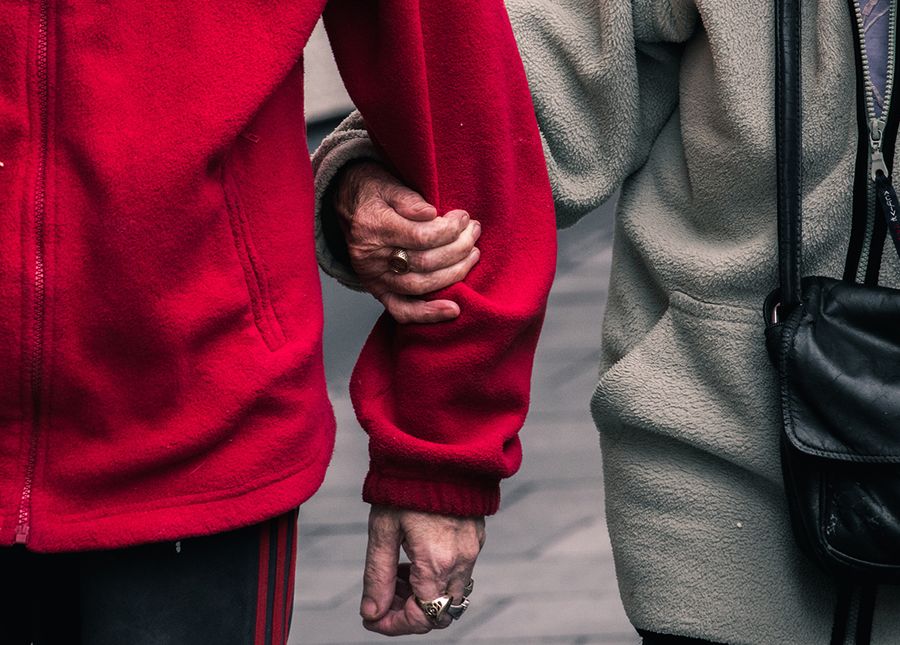 Residential aged care- cropped photo of an older couple's torsos walking, with arms linked, One is wearing a red jacket and the other grey with a black shoulder bag