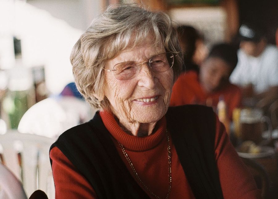 Photo of an older lady, sitting at a cafe while in respite, wearing a rust coloured polo neck with black vest smiling at someone off camera
