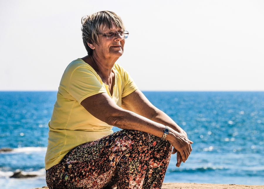 Caring for someone with dementia- Older lady sitting smiling at the beach in a yellow shirt