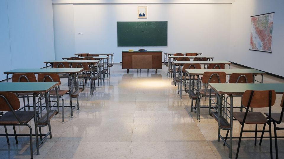 A picture down the middle of a classroom looking at a large desk and blackboard with rows of desks on either side.