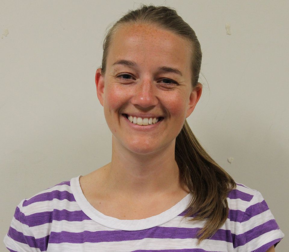 A headshot of Stacey with a big smile wearing a white and purple striped shirt in front of a grey background.