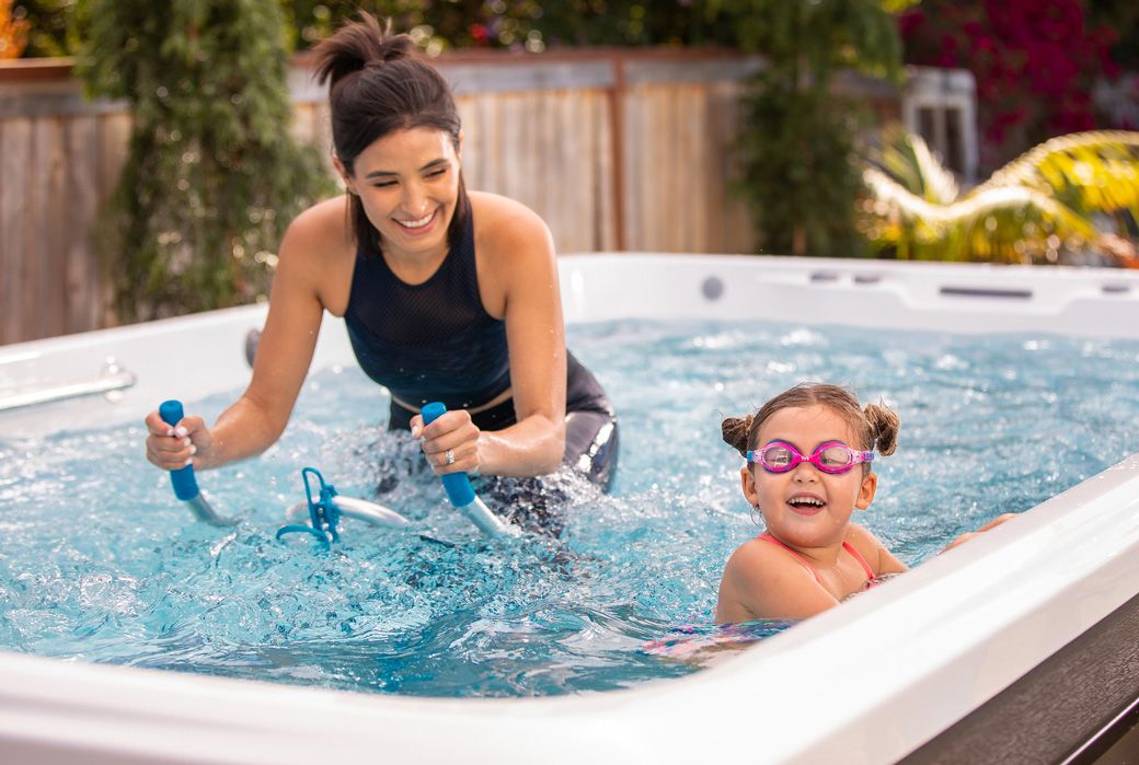 A picture of a smiling woman aqua cycling and watching her daughter as she plays in their swim spa.