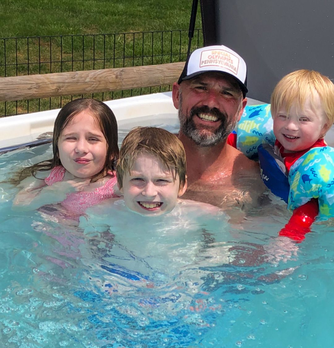 A picture of a man surrounded by his three children in their family pool, all smiling.