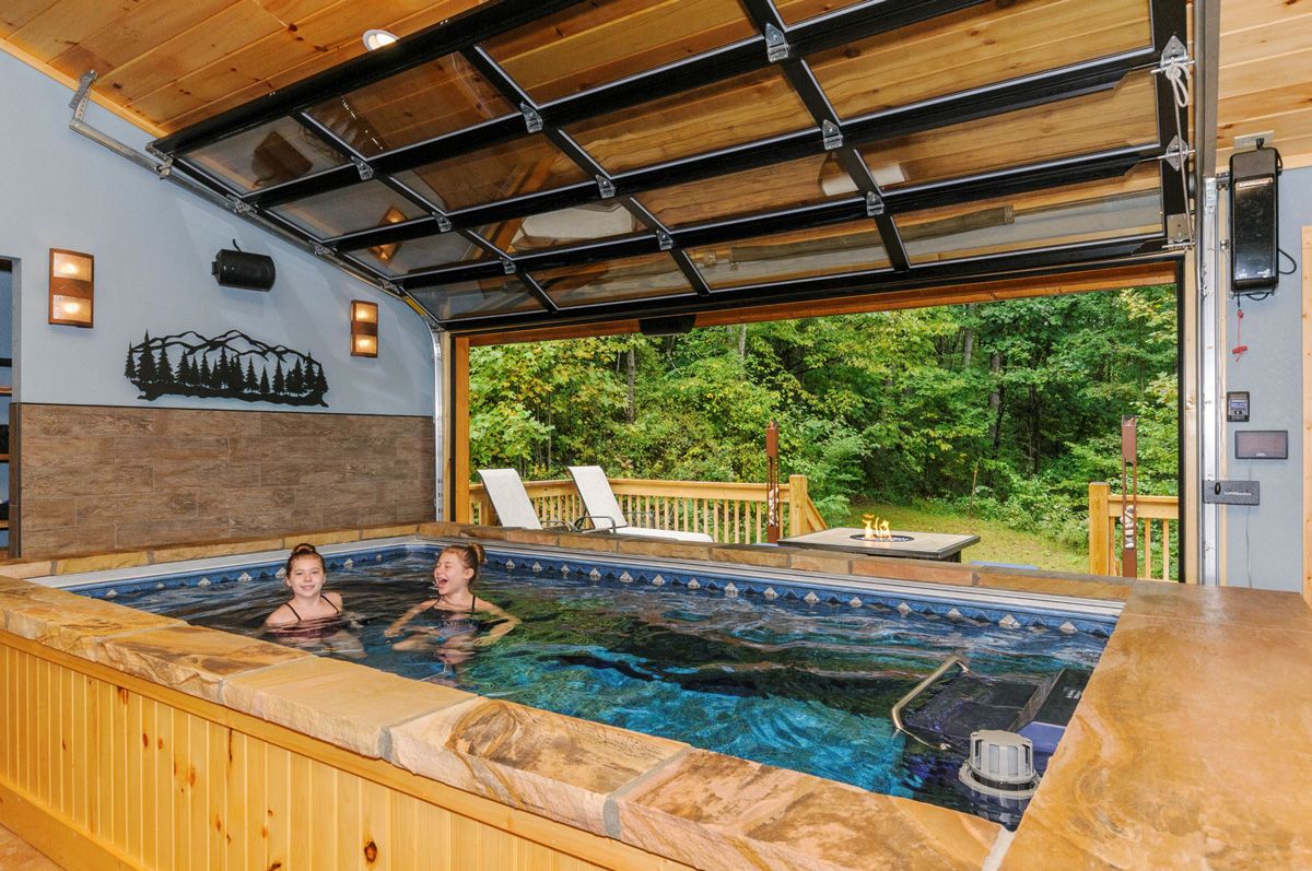 A picture of two girls smiling and swimming in an indoor garage pool with the garage door open to reveal a fire-pit and seating area.
