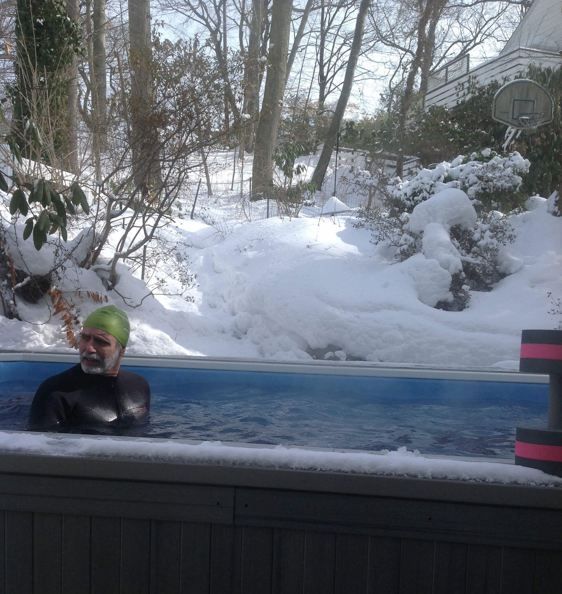 A picture of a man in a wetsuit and swim cap standing in an outdoor pool in the snow.
