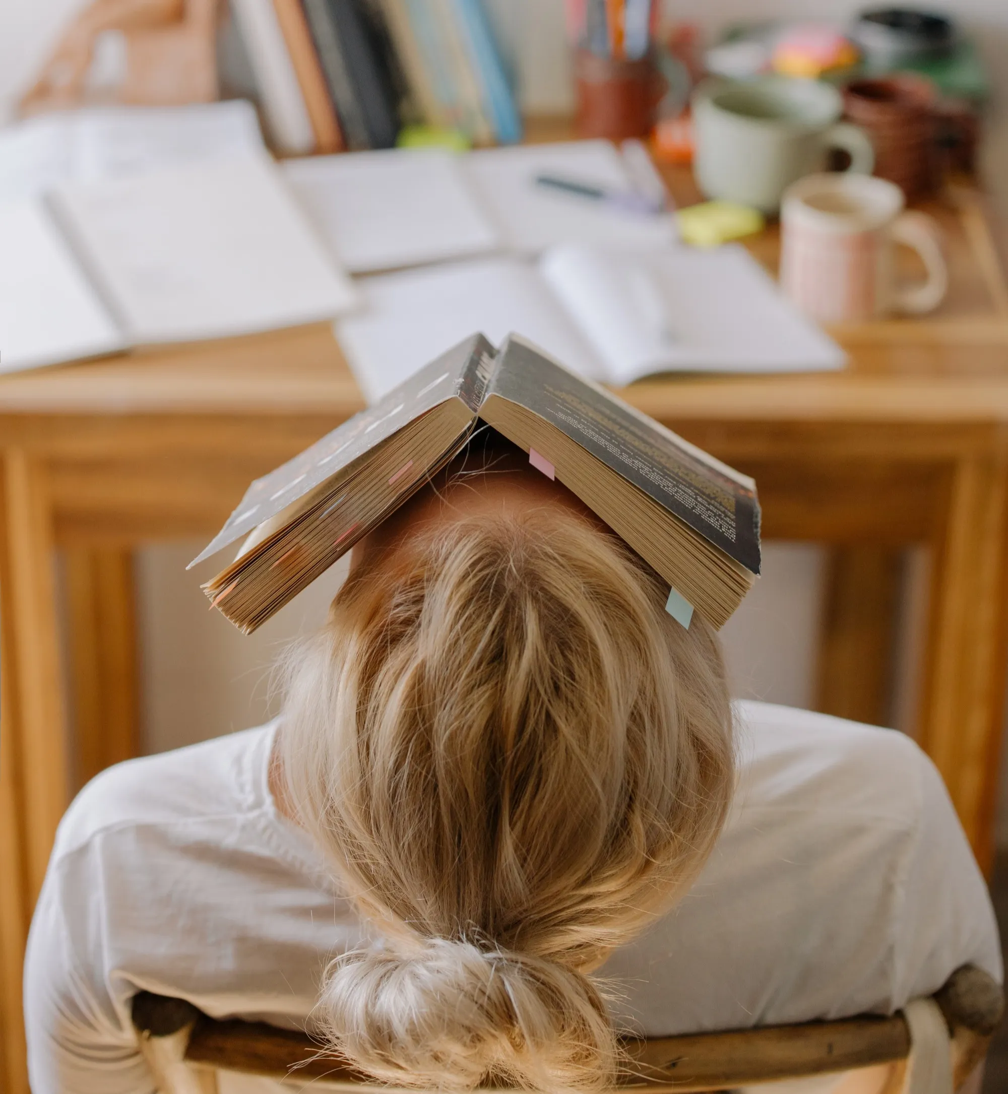 A woman leaning backwards on her chair with a book her head. There is a lot of work in front of her in terms of books and files, she seems totally done.