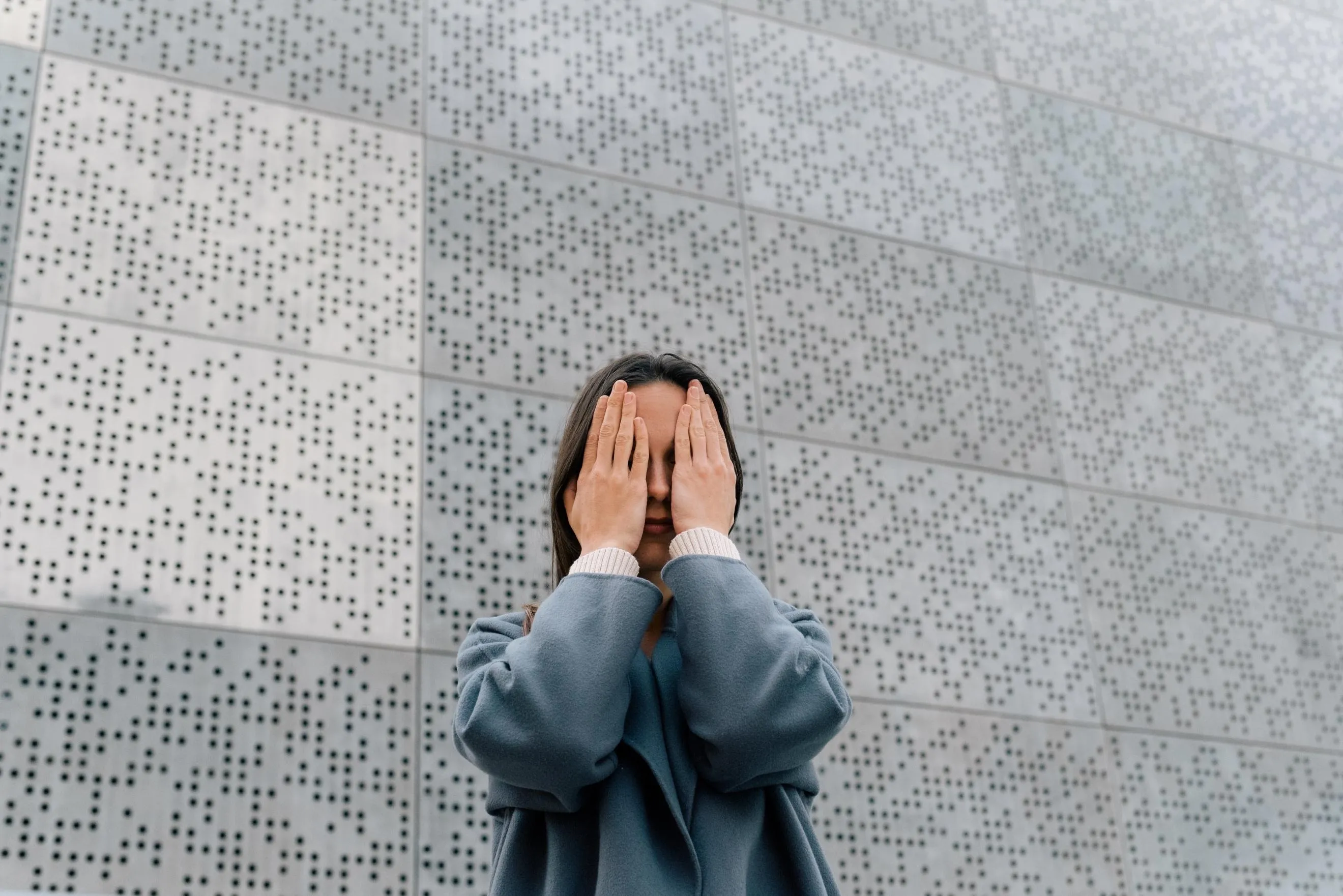 A girl closing her eyes with her hands, standing in front of a grey wall covered in braille markings