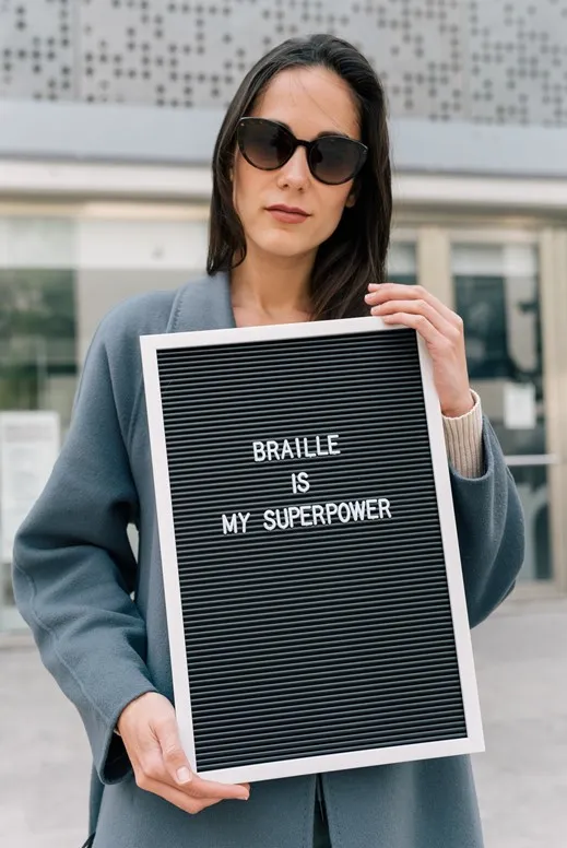 Vrouw met zonnebril die een bord omhoog houd met daarop de tekst: Braille is my superpower