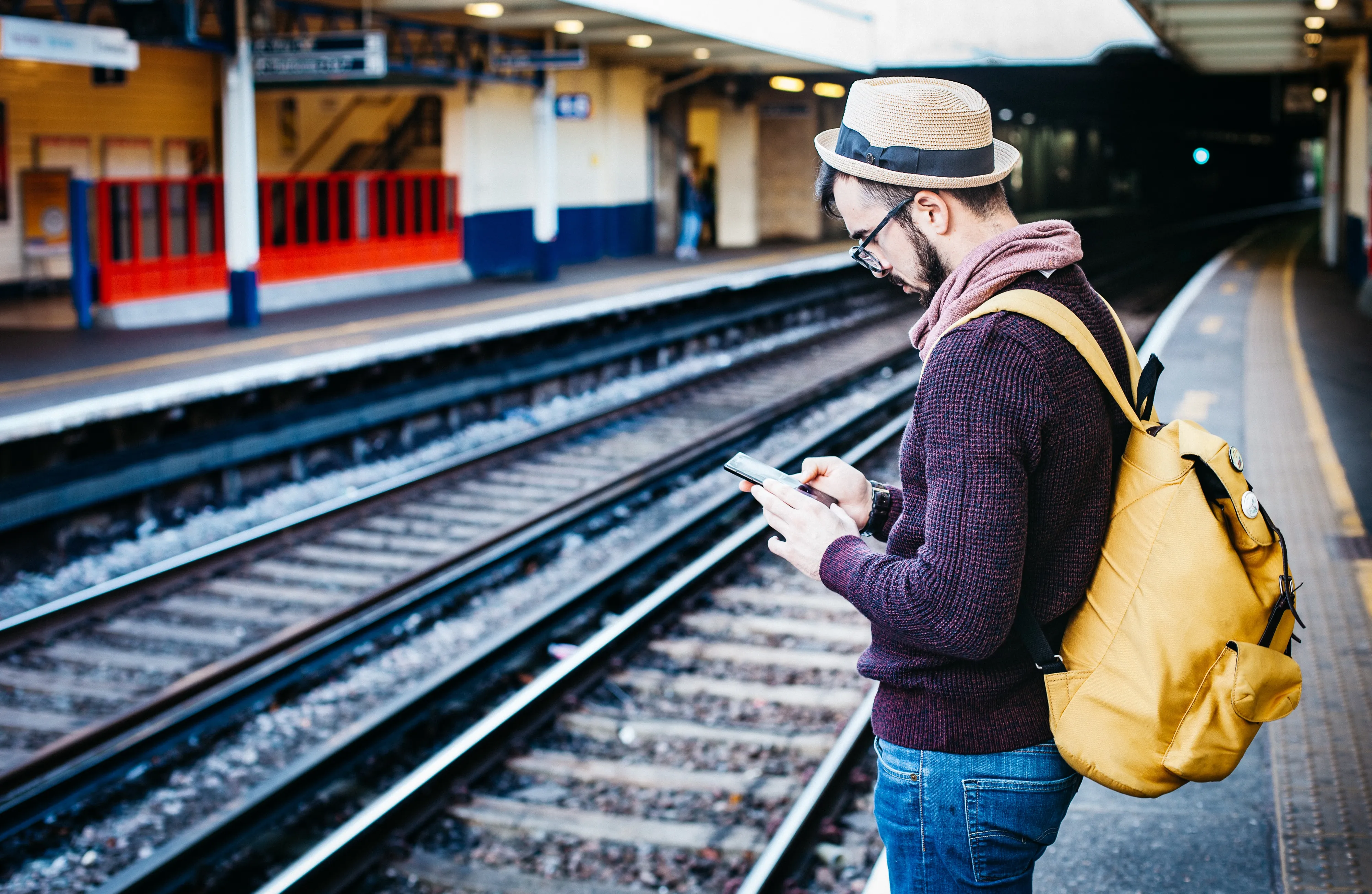 Man standing close to a track at a train station holding his smartphone and wearing a bright yellow bag.