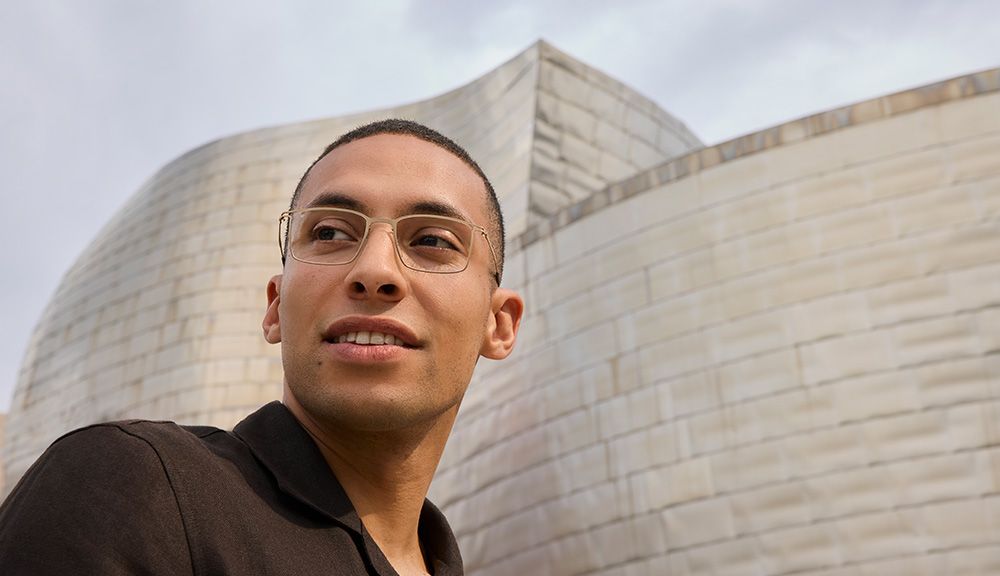 Portrait of a man wearing Titanium Reframed titanium eyeglasses with a sculpted full-rim frame, standing outdoors in front of the Guggenheim Bilbao Museum.