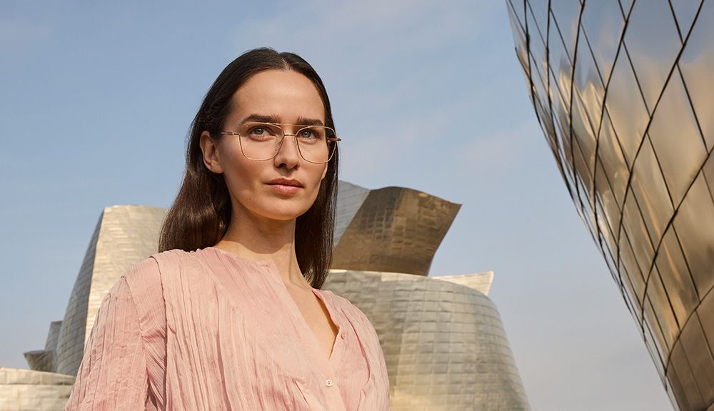 Portrait of a woman wearing Pure Visionary titanium eyeglasses with an ultra-thin gold profile wire frame, standing outdoors in front of the Guggenheim Bilbao Museum.