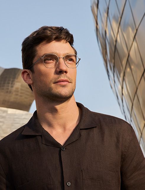 Portrait of a man wearing Pure Visionary titanium eyeglasses with an ultra-thin profile wire frame, standing outdoors beside the Guggenheim Bilbao Museum.