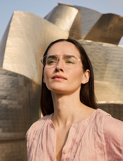 Portrait of a woman wearing Pure Visionary titanium eyeglasses with an ultra-thin gold profile wire frame, standing outdoors in front of the Guggenheim Bilbao Museum.