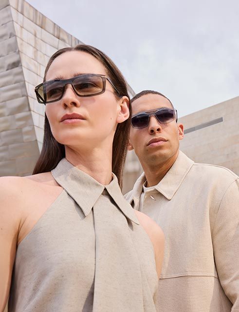Two models wearing modern, lightweight sunglasses stand in soft sunlight against a sculptural architectural backdrop, conveying confidence and effortless summer style.