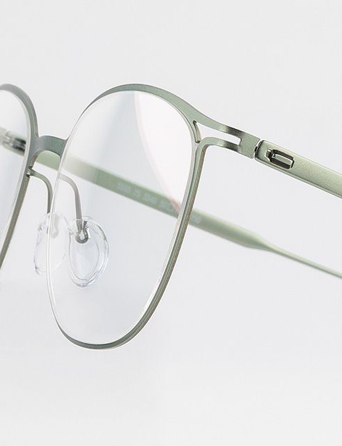 Close-up of Titanium Reframed eyeglasses showing the laser-cut titanium full-rim frame, flat sculpted profile, and minimalist snap hinge in a soft green finish.