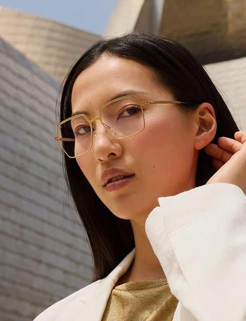Portrait of a woman wearing Titanium Reframed titanium eyeglasses with a sculpted full-rim frame, standing outdoors in front of the Guggenheim Bilbao Museum.