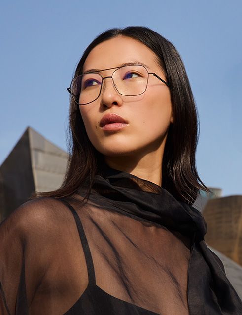 Portrait of a woman wearing Pure Visionary titanium eyeglasses with an ultra-thin profile wire frame, standing outdoors in front of the Guggenheim Bilbao Museum.