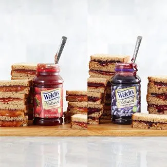 Two jars or Welch's Natural Strawberry and Grape Spead surrounded by stacks of peanut butter and jelly.