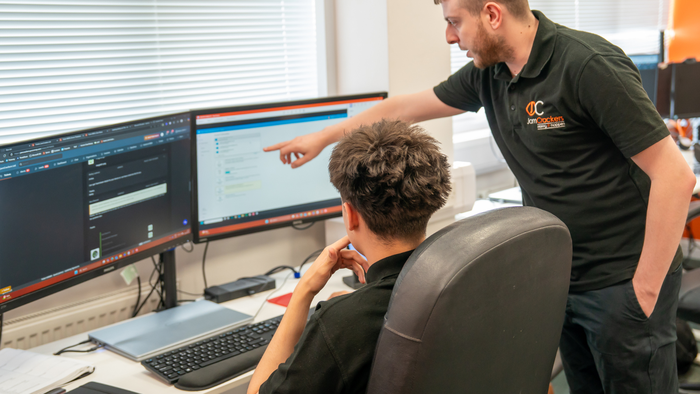 Two men in a tech office setting, one seated and one standing, discussing content on dual computer monitors.