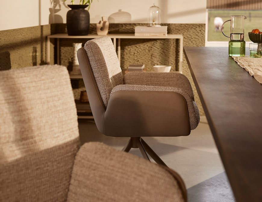 Modern dining chair with textured upholstery beside a wooden table in a sunlit room with a bookshelf and decorative items in the background.
