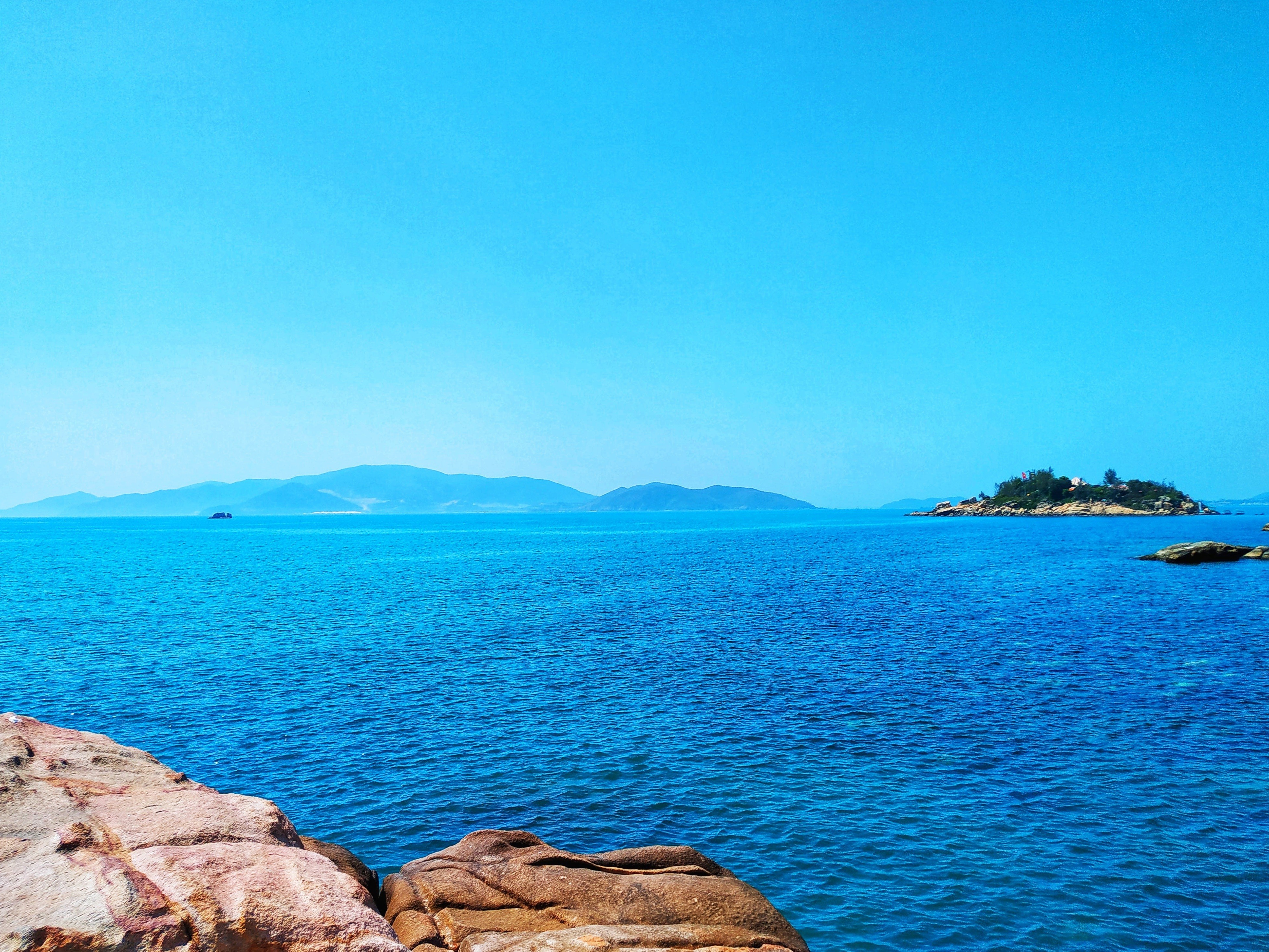 Alt Text Clear blue sea with distant islands on the horizon, rocks in the foreground, and a bright blue sky above.