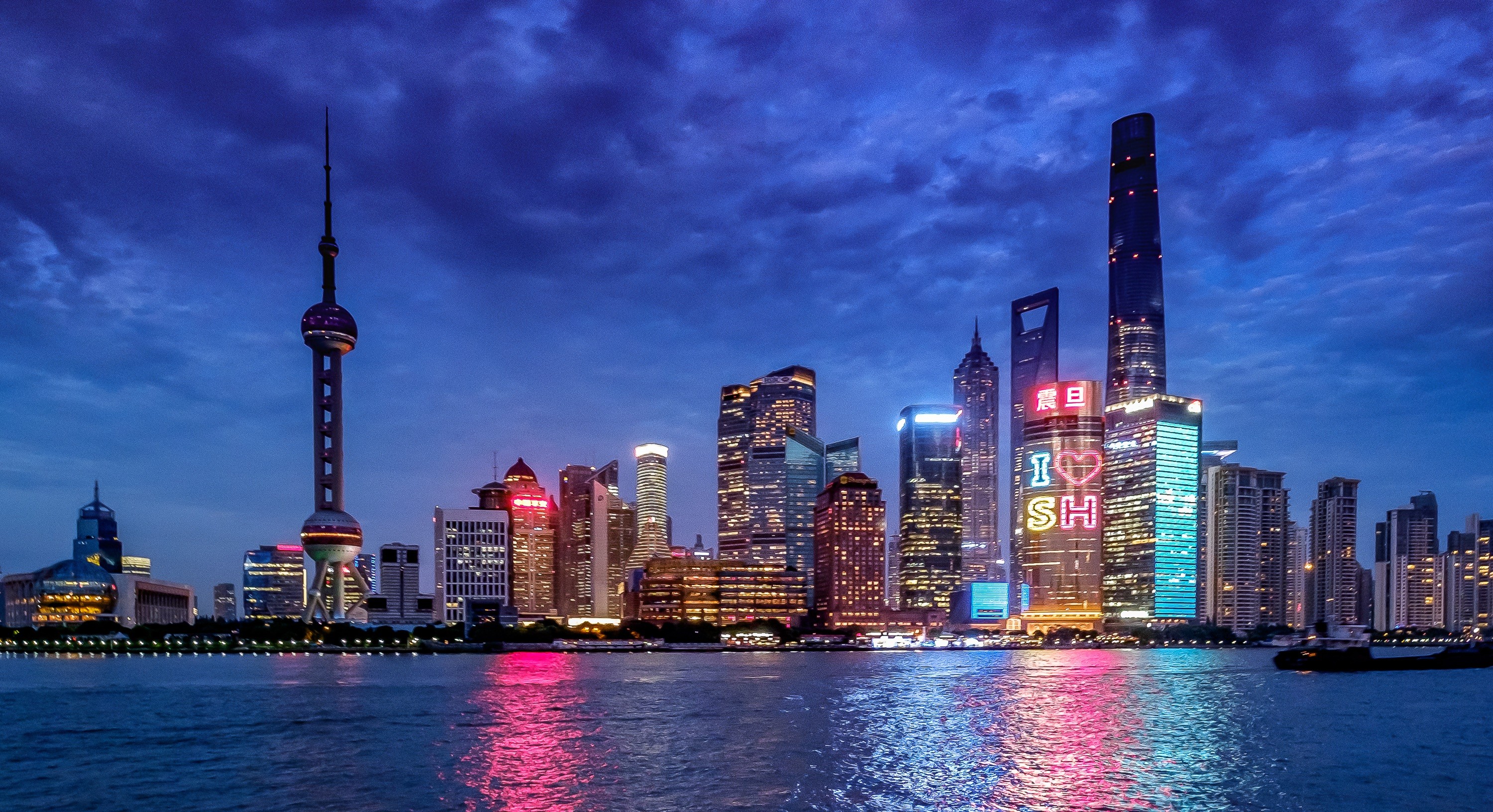 Shanghai skyline at night with illuminated skyscrapers, the Oriental Pearl Tower, and colorful reflections on the Huangpu River under a cloudy sky.