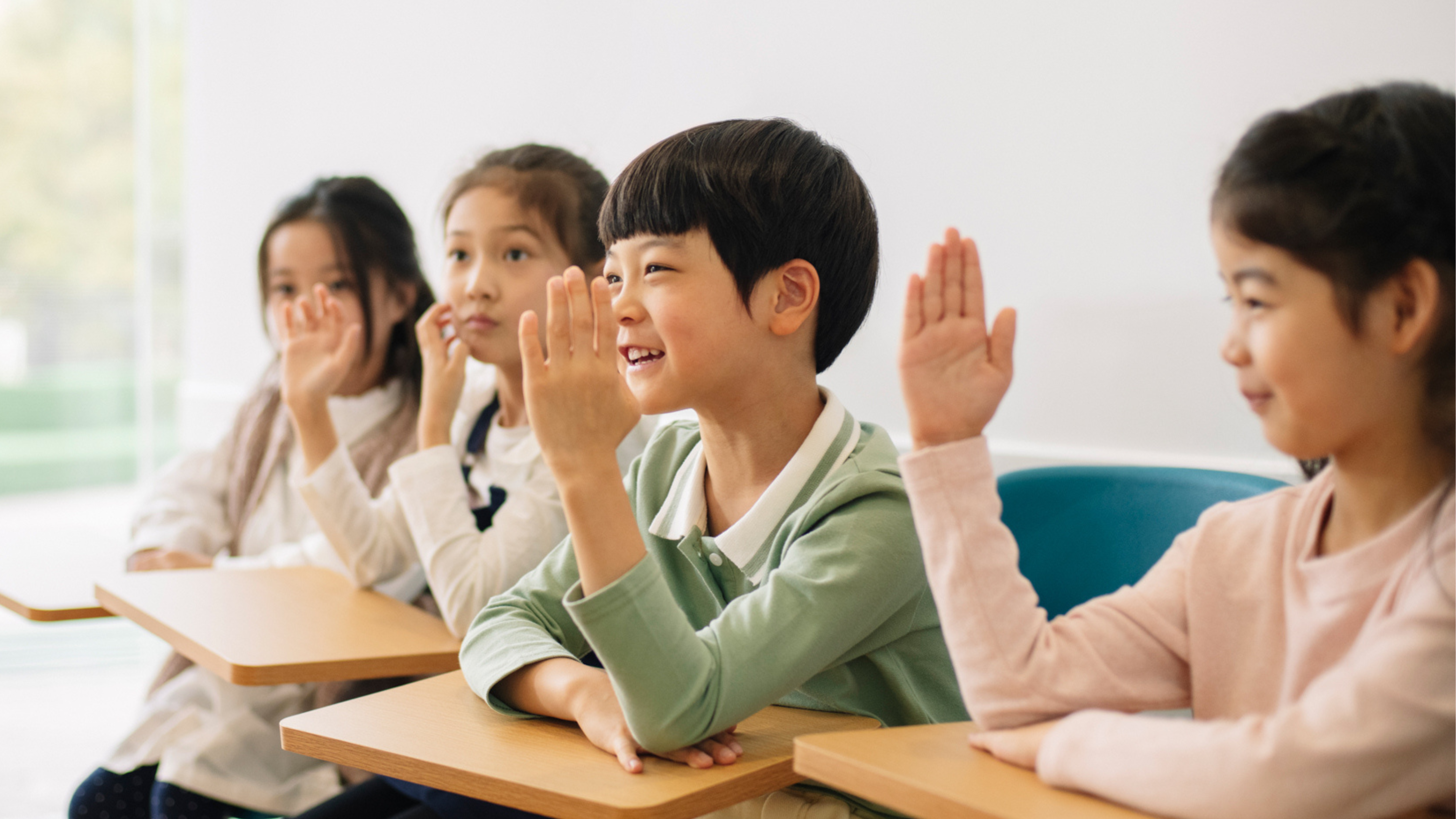 Four children seated at desks in a classroom, eagerly raising their hands, with focused expressions on their faces.