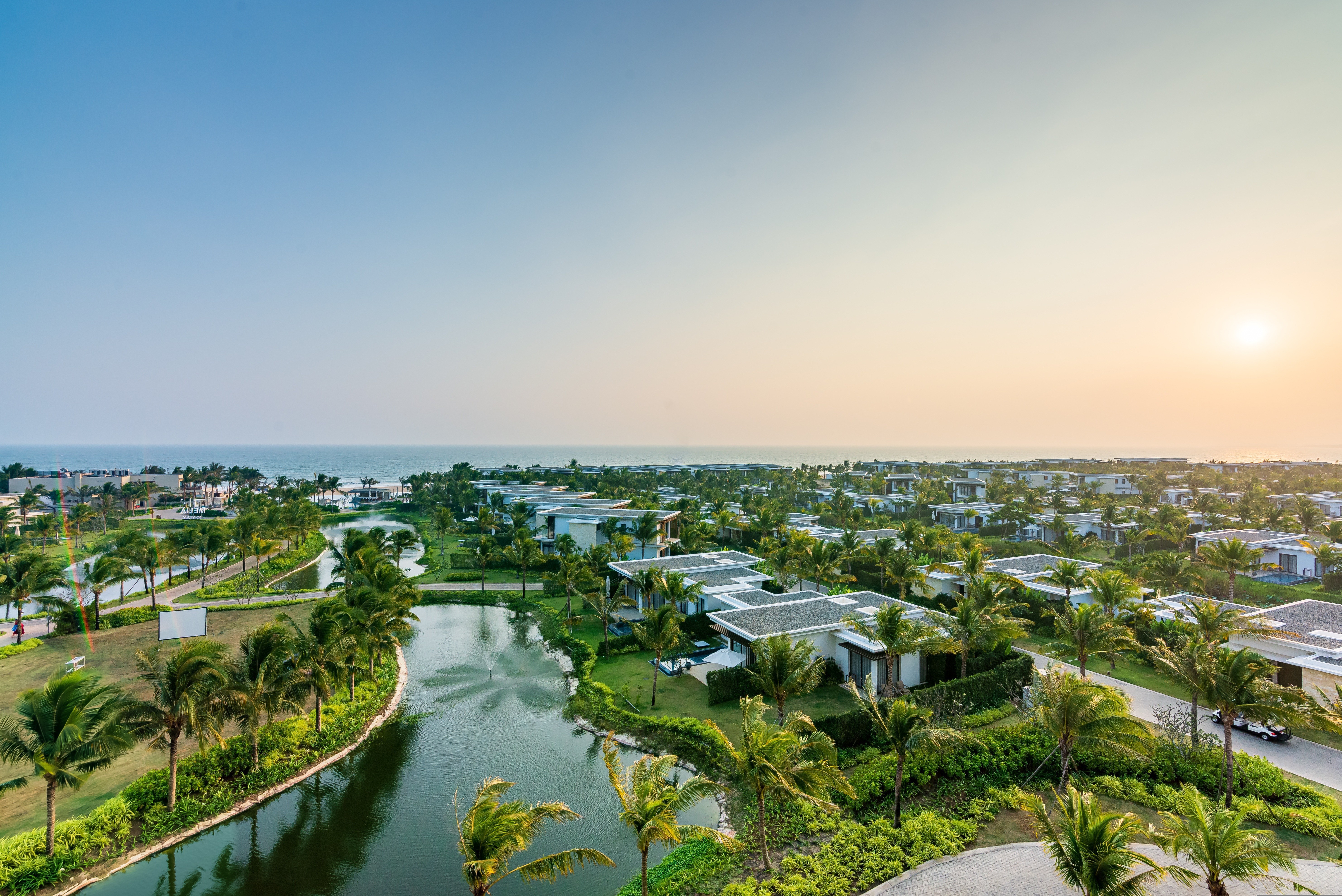 A scenic view of a river with lush green trees, surrounding buildings under construction, and vehicles parked on a road beside the water.