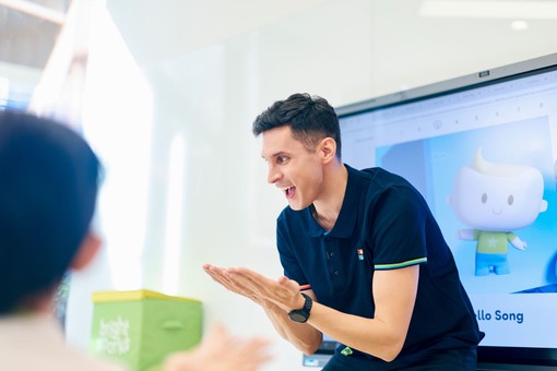 Smiling man in a navy polo engages with a child, clapping hands in a classroom setting, with a cartoon character displayed on the screen behind