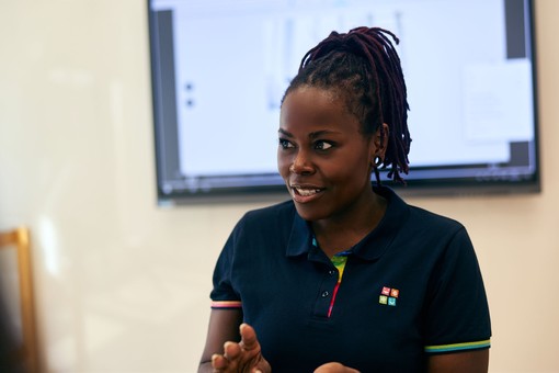 Smiling woman in a navy polo engages with a child, clapping hands in a classroom setting, with a cartoon character displayed on the screen behind