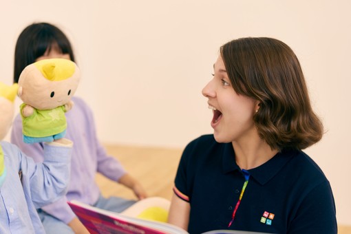 A woman with short brown hair smiles, holding a book, while a child holds a cute plush toy with a yellow tuft, in a playful setting.
