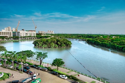 A scenic view of a river with lush green trees, surrounding buildings under construction, and vehicles parked on a road beside the water.