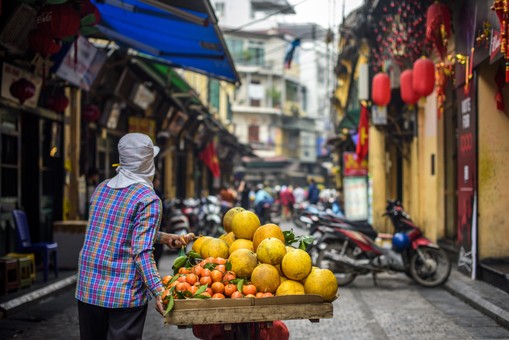 A person in a plaid shirt and covered head pushes a cart with pomelos and oranges through a narrow street, surrounded by motorcycles and buildings.