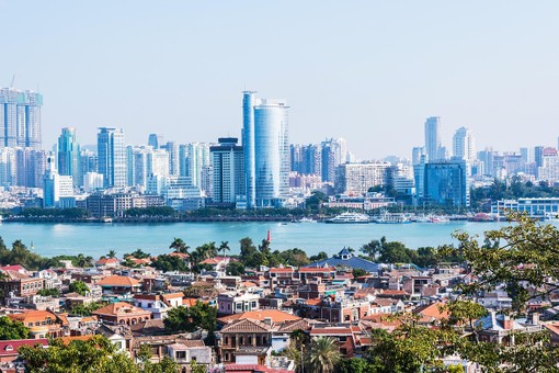 The image shows a cityscape with modern skyscrapers across a river, traditional red-roofed buildings in the foreground, and greenery on a sunny day.